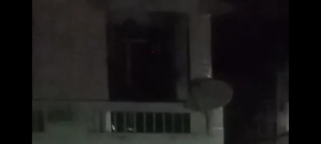 Frame de Modo Noturno (Calebe Lopes, 2020). Uma imagem consideravelmente pixelada  com foco na sacada de um apartamento, da qual é possível visualizar os olhos de uma figura humanóide, com a cabeça dela em destaque pelo escuro da madrugada.